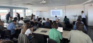 Photo from the back of the conference room with all the participants looking at the screen and the presenter